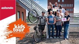 Team von astendo steht auf Treppe, hält Schilder mit "kids-Tour" hoch, daneben ein Kollege mit Fahrradhelm und Fahrrad